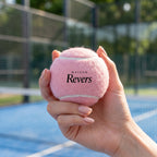 Main manucurée tenant une balle de padel rose Maison Revers sur un terrain