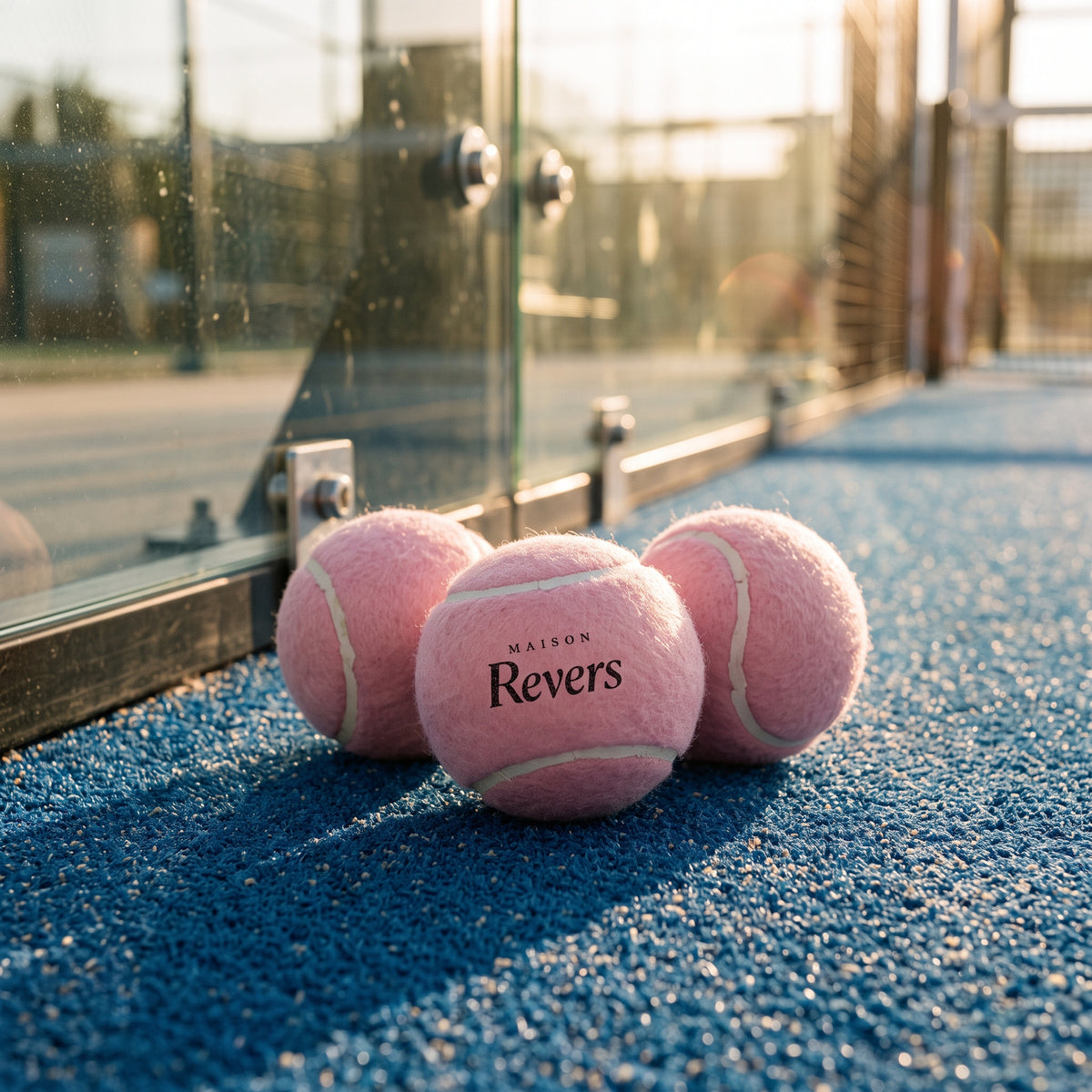 Trois balles de padel roses Maison Revers au pied de la vitre d'un terrain
